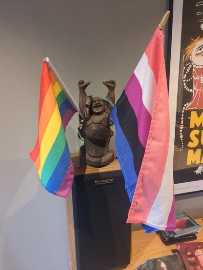 Two flags, a rainbow flag and a gender-fluid flag, are displayed on a light wooden speaker, with a statue of a happy Buddha in the middle holding onto the flags!
