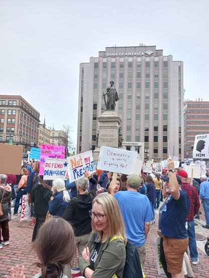 A city square with people holding up signs. There is still space between groups of people. The people are surrounding a statue of the goddess Nike ("Our Lady of Victories"). In the distance is a grey, tall office building with the words, "Bank of America" on the top level. Some of the signs from the protest read: "Defend Democracy," "Not My President," "Stop him," and "Democracy is worth fighting for." The sky is grey.
