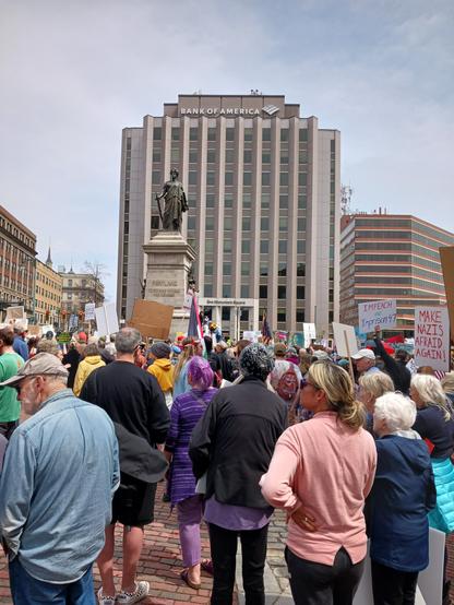 A city square with people holding up signs. The streets are crowded. The people are surrounding a statue of the goddess Nike ("Our Lady of Victories"). In the distance is a grey, tall office building with the words, "Bank of America" on the top level. There are Pride flags, Palestinian flags, and a signs that read: "Impeach the President" and "Make Nazis Afraid Again." The sky is cloudy, but blue sky is starting to appear.