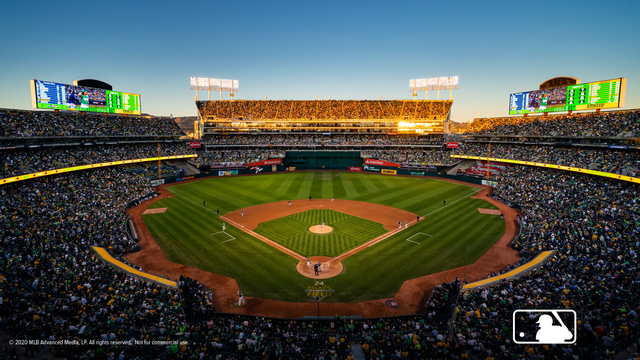 The former home of the Oakland Athletics, the Oakland Coliseum, during a baseball game. The stands are full and the field gleams in the golden hour near sunset.