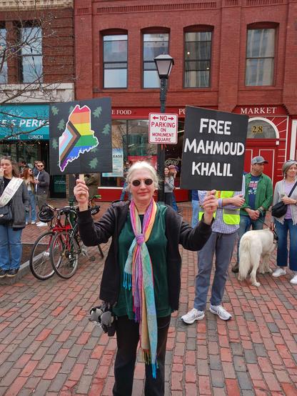 A person wearing green and black and a pastel rainbow scarf. They have white hair and are wearing dark oval sunglasses, and holding up two signs: One sign is an image of the state of Maine with the inclusive Pride flag within the borders; the second sign reads, "Free Mahmoud Khalil".