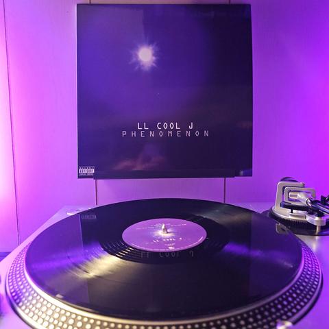 A vinyl record sits on a turntable. Behind the turntable, a vinyl album outer sleeve is displayed. The front cover a glowing white light against a dark purple background with the text "LL COOL J PHENOMENON" and a parental advisory label at the bottom left.