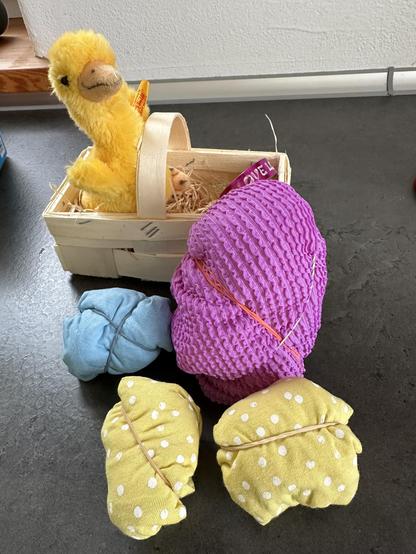 kleines Holzkörbchen mit Holzwolle, aus dem ein Steiff-Küken herausschaut. Davor liegen bunte kleine Textilien, mithilfe von Gummibänden in Eierform gebracht.