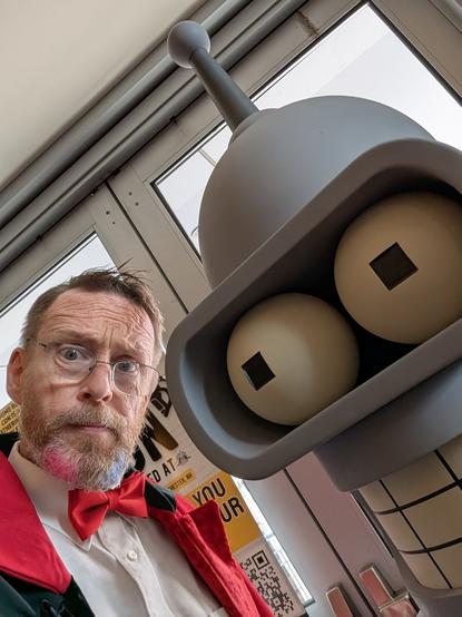 A man cosplaying as Harley Quinn standing next to a Bender statue from Futurama.