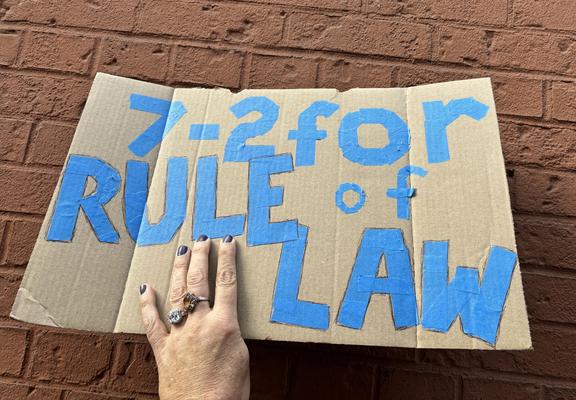 Me holding my new 2-sided protest sign, up against the painted brick wall of Port Authority. In blue painter’s tape it says “7-2 for rule of law”. My left hand nails are painted purple.