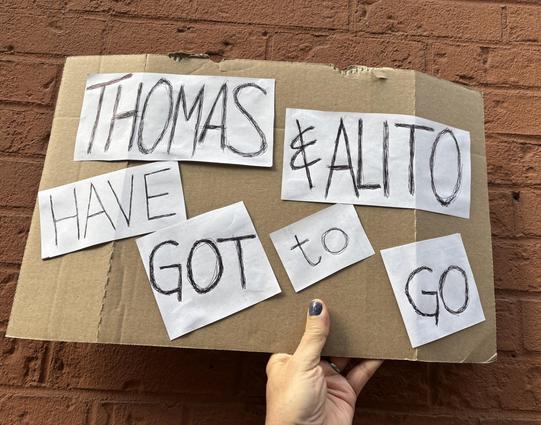 Me, holding up the other side against the painted brick wall outside Port Authority. There are white rectangles and squares, each with words: “Thomas & Alito have got to go”