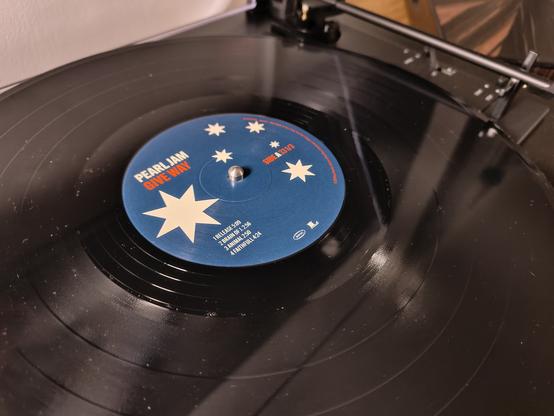 Das Bild zeigt ein Plattenspieler mit einem schwarzen Vinyl-Album, das auf dem Plattenspieler platziert ist. Im Zentrum des Albums befindet sich ein blauer Etikett mit dem Text "PEARL JAM GIVE WAY" in weißer und roter Schrift. Das Etikett ist mit weißen Sternen verziert. Der Plattenspieler hat einen Arm, der über das Album schwebt, bereit, die Platte zu spielen. Der Hintergrund ist unscharf, wodurch der Fokus auf dem Album und dem Plattenspieler liegt.