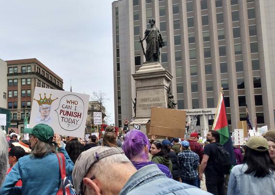 Protestors gathered around a statue of a woman with a sword (Our Lady of Victories) in a city square. One of the protestors is holding a Palestinian flag, and another holds a sign that depicts Trump as a king, saying, "Who can I punish today?"