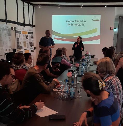 Zu sehen ist ein langer Tisch, an dem Menschen sitzen, die einer Frau mit Mikrofon und einem Mann zuhören. Auf einer Leinwand steht neben dem Logo der nestbau AG "Guten Abend in Münnerstadt "