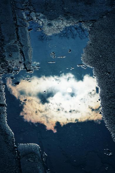 Das Bild zeigt eine Pfütze, die sich auf einer Asphaltstraße befindet. In der Pfütze spiegelt sich der Himmel, der von Wolken und der Sonne geprägt ist, wodurch eine schöne Verbindung zwischen Himmel und Erde entsteht. Der Asphalt rund um die Pfütze ist uneben und hat Risse, die die Textur des Bodens hervorheben. Die dunkle Farbe des Asphalts kontrastiert mit der helleren Spiegelung des Himmels in der Pfütze, was einen interessanten visuellen Effekt erzeugt.