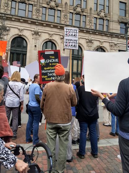 A crowd of people in front of a stylish old building, holding signs. One man, wearing brown and green-green clothes and wearing an orange cap, holds a sign that reads: "This GNOME is more qualified for Homeland Security than Kristi Noem". The sign has an illustration of a gnome wearing brown and green and an orange cap.