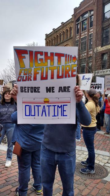 A protestor holding up a sign that reads: 
FIGHT FOR OUR FUTURE!  (In "Back to the Future letters")
BEFORE WE ARE
OUTATIME (which looks like a license plate).