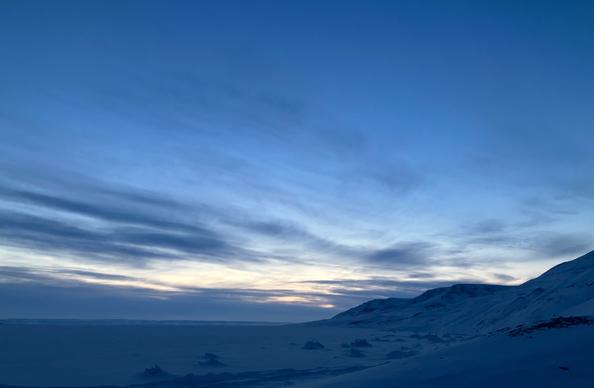 A light blue midnight school with wispy clouds. The horizon is bright with a hint of orange. The mountain and ocean are covered in snow and shadow.