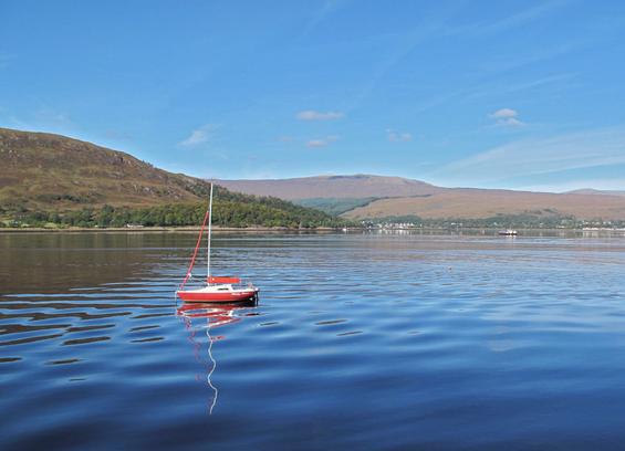 A red sailboat floats in calm blue water, surrounded by green hills and a clear sky. The reflection of the boat and the surrounding landscape is visible on the water's surface.