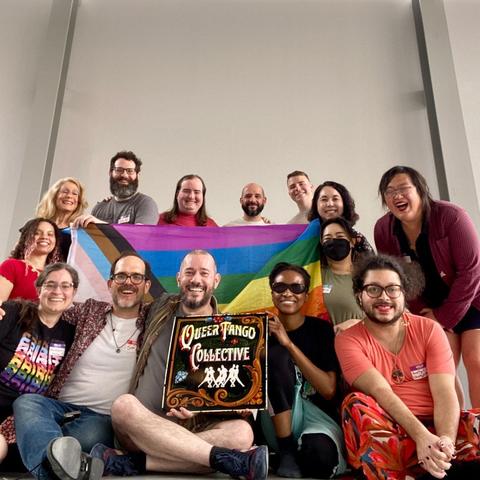 14 people smile around a progress pride flag and Queer Tango Collective sign. They are of various races and cover practically every stage of adulthood.
