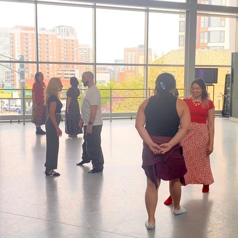 Three couples practice having one person mirror the other in walking forward and back and changing weight without touching. The nearest couple, we see a person with long black hair in a claw clip, a black sleeveless top, and a red flannel tied around the waist, dancing with someone else in a red shirt and red and white skirt who has brown and pink curly hair.