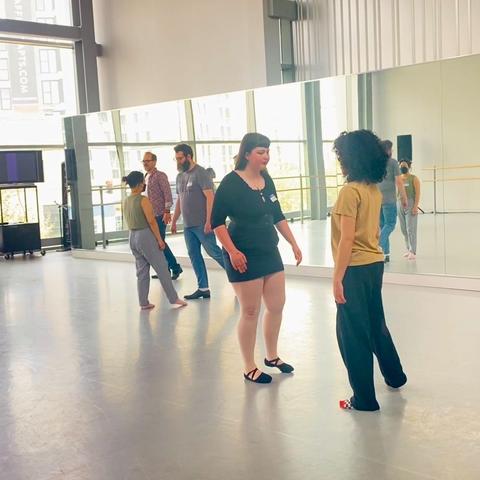 Two couples, without touching, practice mirroring each other, legs outstretched as one half of each couple leads the other to step. The instructor is in the background. The nearest couple includes a person with big curly hair, a yellow shirt, and black pants along with another person with long straight hair, a black shirt and waist cincher, and short black skirt along with black ballet slippers.