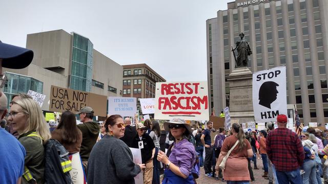 Protestors in a city square holding up signs. Some signs read: USA NOT FOR $ALE!, RESIST FASCISM! and STOP HIM!