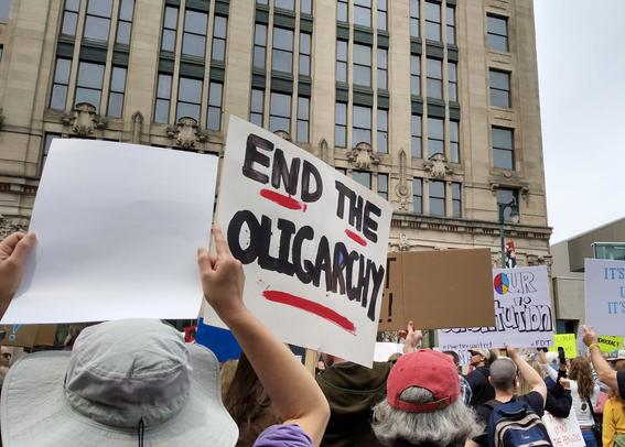 A crowd of people holding up signs. One sign reads, END THE OLIGARCHY! In big black letters with red underlines.