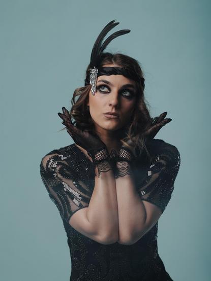 The image shows a person dressed in an outfit reminiscent of the 1920s flapper style. They are wearing a black sequined dress with sheer sleeves, a headband adorned with feathers and a jeweled brooch, and fishnet gloves. Their pose is expressive, with hands near their face, and they have bold eye makeup.