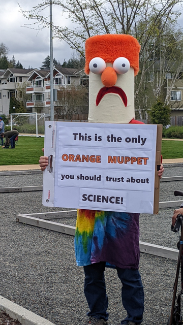 The image shows a person holding a protest sign in a costume of Beaker from the Muppets. The sign says "This is the only ORANGE MUPPET you should trust about SCIENCE!"