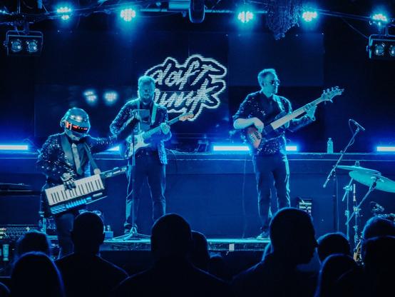 Photo of three band members on a stage playing live music. The lead has a keytar, the two others have electric guitars. In the background, the Daft Punk logo is shown on a screen.