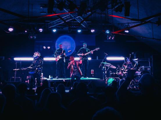 Five band members on a stage with the audience’s heads in the foreground. Centre is the main singer with a red leather jacket and matching red hat.