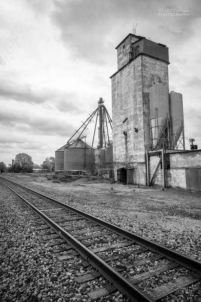 Picture of an old concrete grain silo next to multiple short, round, metal grain storage silos.  A railroad track runs across the foreground.
