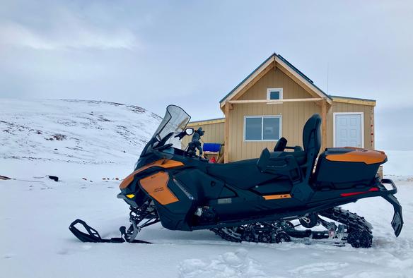 An orange and black snowmobile parked in front of a cabin in a snowy landscape with overcast skies.