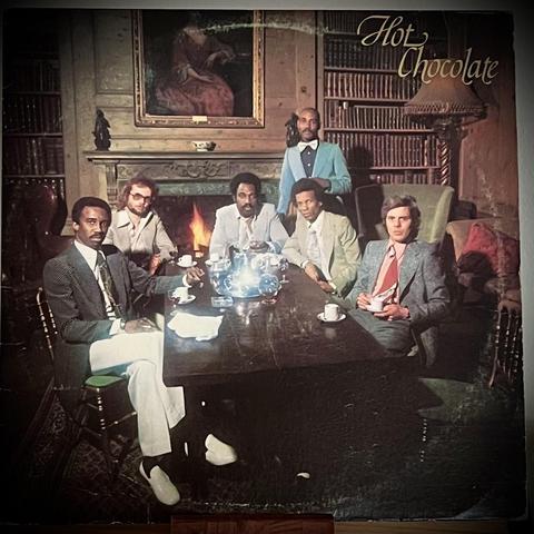 A record cover featuring a group of six men is seated around a large table in a richly decorated room with bookshelves and a fireplace. They are dressed in formal attire and sipping from cups, with a teapot and cups arranged on the table. The scene has a vintage feel.