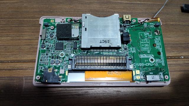 A primeira imagem mostra a placa-mãe do Nintendo DS Lite com o slot de cartuchos, conectores e o processador visíveis. A segunda imagem exibe a tela superior com a fita flat e fios soldados — possivelmente para modificação de iluminação ou som. A terceira imagem apresenta a carcaça traseira com os alto-falantes e a tela ainda montada. Esse tipo de desmontagem é comum em projetos de reparo ou modding.

English:
The first image shows the Nintendo DS Lite motherboard, including the cartridge slot, connectors, and visible processor. The second image displays the top screen with its flex cable and soldered wires — likely for a mod or backlight/sound tweak. The third image shows the back shell with the speakers and screen still attached. This type of disassembly is common in repair or modding projects.