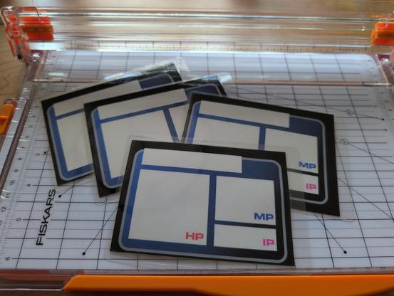 A set of four laminated cards sitting on a paper cutter. Each card has spaces for writing "HP", "MP", and "IP."