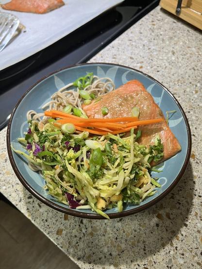 A blue floral bowl holds soba noodles, a baked salmon fillet, and broccoli slaw salad, garnished with pickled carrots and sliced green onions.