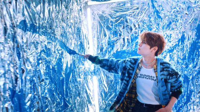 Seungmin wielding a paintbrush in a room covered in crinkled foil. He's wearing a fuzzy cardigan and a plaid shirt over a t-shirt that says SUNDAY FRUIT TOUR
