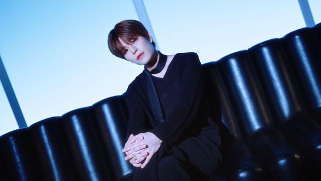 Seungmin sitting on a shiny black couch with his legs crossed and his hands clasped over his knee. He has his head tilted to one side and is judging you thoughtfully. His all-black outfit is quite simple but also regal, and he has a scarf around his neck that looks a bit like a collar