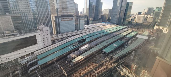 A wide-angle daytime view of Tokyo Station, seen from a high vantage point. The image shows the station’s platforms with their characteristic green roofs, multiple train tracks, and surrounding modern skyscrapers. The architectural contrast between the old red-brick Tokyo Station building in the foreground and the tall glass office towers in the background is clearly visible.