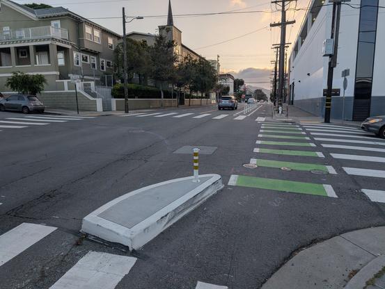 Looking west along the bike lane on 17th St at San Bruno Ave, across the odd droplet-shaped curb installed in a rather baffling attempt at emulating a protected intersection.  The bike lane is unpainted on either side of the intersection, but dashed-green-striped inside it, failing at continuity and instead throwing a visual transition right behind the hazard the curb presents to cyclists not expecting the bike lane to abruptly shift 2-3 feet to the right.  Rubber tire marks along the inside of the curb suggest cyclists have already started crashing into it.

It's intended that the curb force drivers to slow more for a sharper right turn that would cross the bike land and crosswalk, reducing their hazard to others and placing more of their arc of turn outside the zone of interference, thus improving their visibility.   However, the curblet has been built extending only a foot or two beyond the San Bruno Ave turn, and is strongly radiused, so it alters cars' path of travel hardly at all.