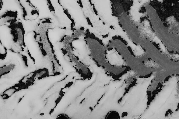 The image shows snow on a surface, creating an interesting pattern with darker areas where the snow has melted or been disturbed. The pattern resembles swirling lines and contrasts between the white snow and the dark surface underneath.