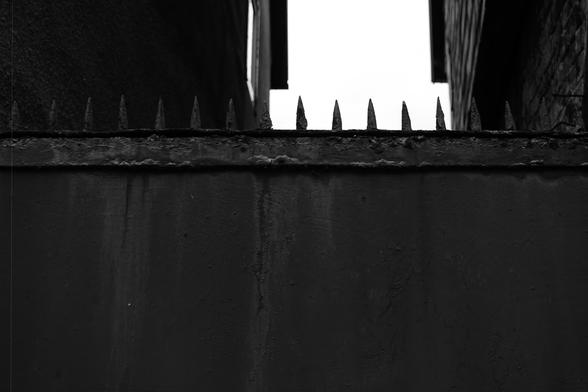 The image shows a black and white photo of a wall topped with sharp spikes. It appears to be an urban setting, with buildings visible in the background. The spikes are designed likely for security purposes, preventing access over the wall. The overall tone is stark and moody.