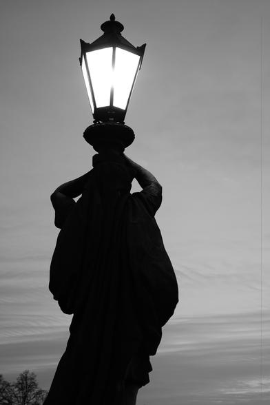 The image shows a black and white silhouette of a statue holding up a street lamp against the sky. The light from the lamp creates a stark contrast with the dark figure of the statue.