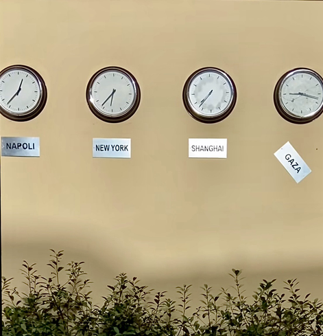 A sand coloured wall in a school yard with clocks hanging on the wall showing the different time in Napoli, Nee York, Shanghai & Gaza - where the time has stopped.