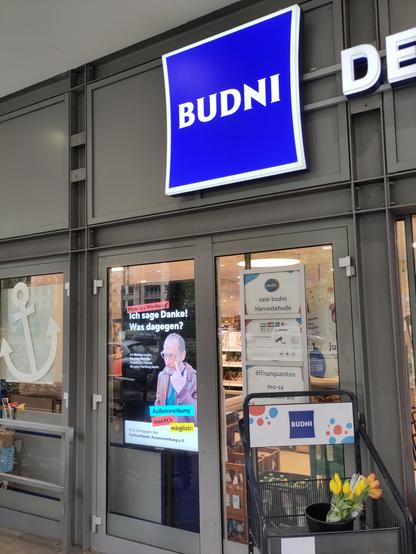 BUDNI. Im Schaufenster prominent ein Werbebildschirm, ca. DIN A1 groß, darauf die Kampagne "Mehr als Werbung. Außenwerbung macht's möglich" des Fachverband Außenwerbung e.V., mit dem Werber, der sich das Augenlid herunterzieht und dem Zitat "Ich sage Danke! Was dagegen?"