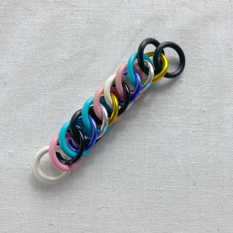 A piece of chainmail with two interlocked sides; one in the colours of the trans pride flag and one in the colours of the nonbinary pride flag.
