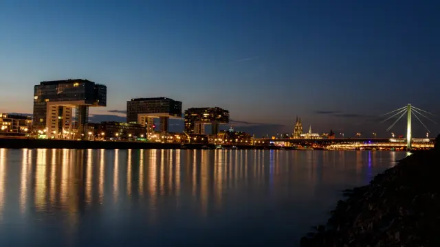 a photograph of the Rheinauhafen (lit. Rheinau harbour) with the three Kranhäuser (lit. Crane Houses) during evening twilight in Cologne. Most of the buildings on the opposite side of the Rhine are already illuminated. The illumination is being reflected in the water. Downstream and on the right side of the picture is the Severins Bridge. In the middle between the last Kranhaus and the bridge is the Cologne Cathedral.