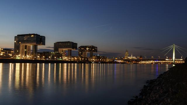a photograph of the Rheinauhafen (lit. Rheinau harbour) with the three Kranhäuser (lit. Crane Houses) during evening twilight in Cologne. Most of the buildings on the opposite side of the Rhine are already illuminated. The illumination is being reflected in the water. Downstream and on the right side of the picture is the Severins Bridge. In the middle between the last Kranhaus and the bridge is the Cologne Cathedral.