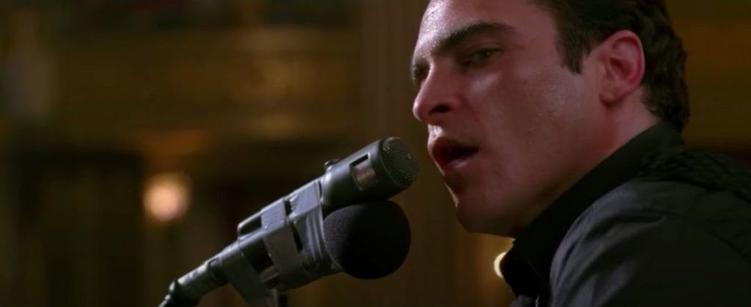 A close shot of a man on a stage, singing into the mic, looking over at someone standing next to him
