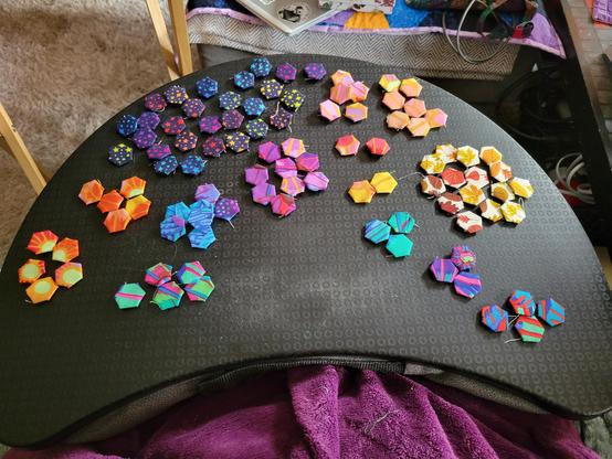 Photograph of maybe 80 half inch hexagons arranged by main color on a black lap tray. Some are dark blue with rainbow stars and many are various cheerful bright colors including peach, teal, blue an green. One small group have leaves on in autumnal colors.