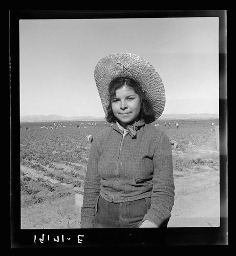 The image is a black and white photograph depicting a young girl standing in an agricultural field. She wears a wide-brimmed straw hat, which suggests she may be protecting herself from the sun while working outdoors. The girl dons a knitted sweater with a scarf around her neck and pants that appear to fit comfortably.

Behind her, rows of crops extend into the distance under a clear sky. It appears to be a sunny day due to the bright light casting shadows on the ground. In the background, several figures can also be seen working in the fields, implying this might be a common activity or setting for these individuals.

The photograph bears text at the bottom left corner with "INZINI - E" which seems like an identifier of some sort. The overall composition suggests that the image is part of a larger documentary work capturing rural life and labor conditions during the time it was taken, possibly in Imperial Valley, California as mentioned in the accompanying information.

This photograph reflects themes such as child labor, agricultural practices, and daily struggles faced by individuals working on farms or fields for economic sustenance.