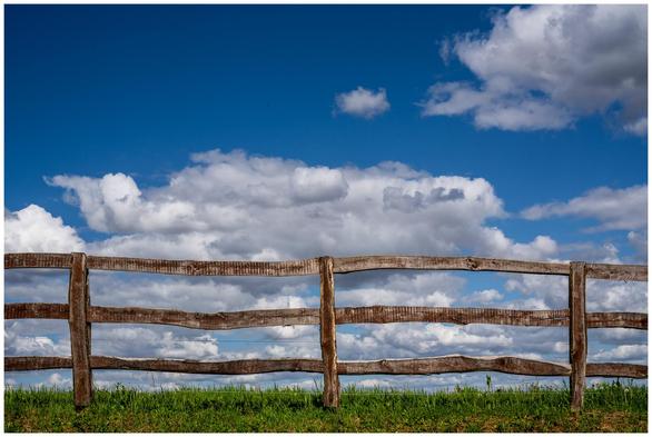 Ranchzaun auf einer bewiesten Böschung gegen Blauhimmel mit Fluffelswolken.
