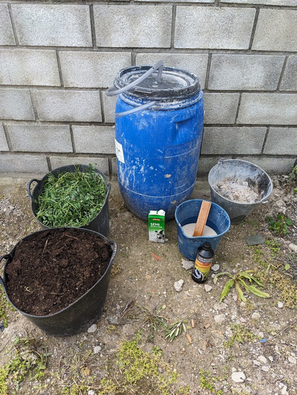 Foto d'un bidó de plàstic gran, envoltat d'una sèrie d'ingredients per a preparar fertilitzant orgànic: un cabàs amb fem, un altre amb herba fresca, un cartró de llet de vaca, un litre de melassa, un poal amb cendra i un poal amb aigua i rent.